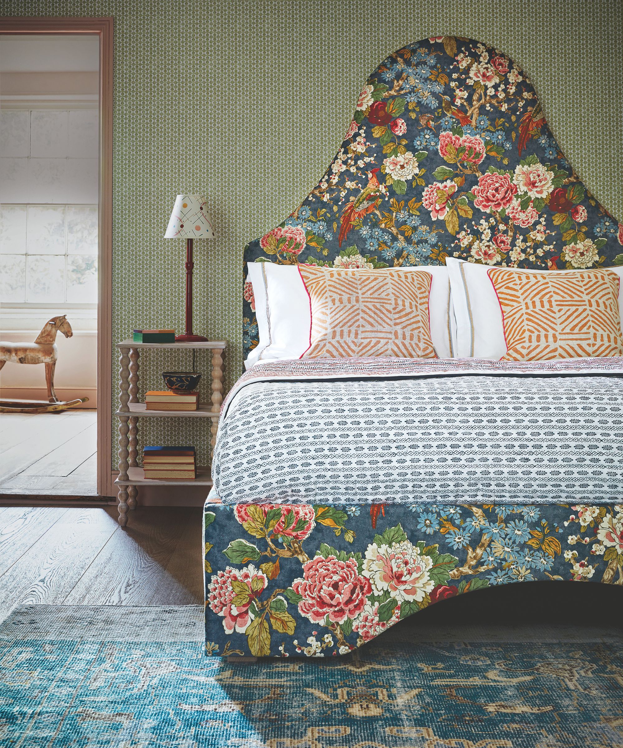 A green maximalist bedroom with a large floral patterned bed with statement headboard, patterned bedding, and orange patterned cushions. Beside the bed is a wooden bedside table with books and a wooden lamp with a white lampshade on top. In the foreground is a blue patterned rug. In the background to the left is an open door with a wooden rocking horse seen beyond.