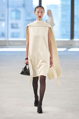 A model wearing a butter yellow scarf satin minidress with black tights and accessories at the S/S 25 Toteme show showcasing the modern socialite trend.