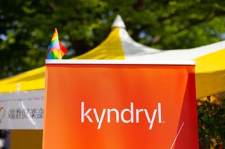 Kyndryl logo in white lettering printed on a poster with an orange background and a rainbow flag attached to the top of it