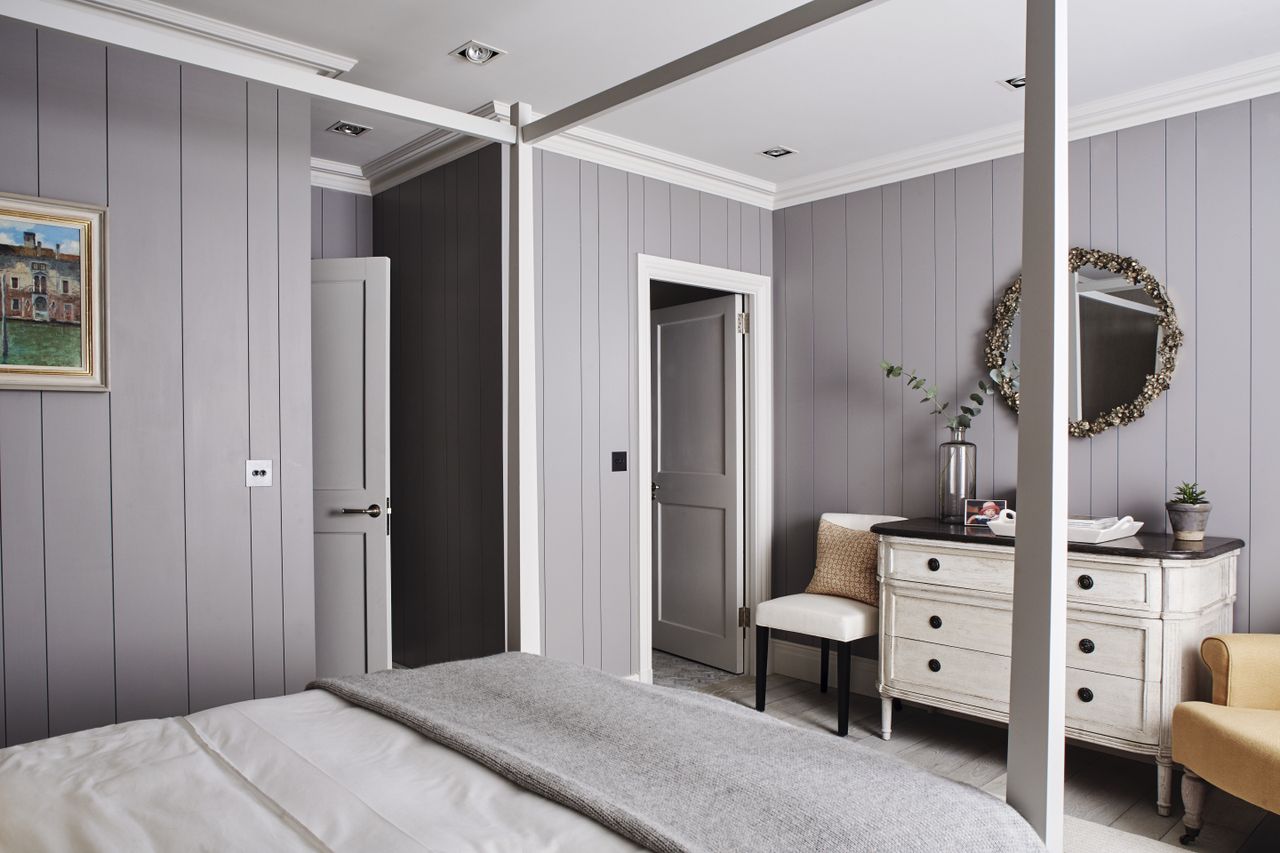 Basement bedroom with four poster bed, grey panelled walls, white painted chest of drawers, and upholstered chairs 