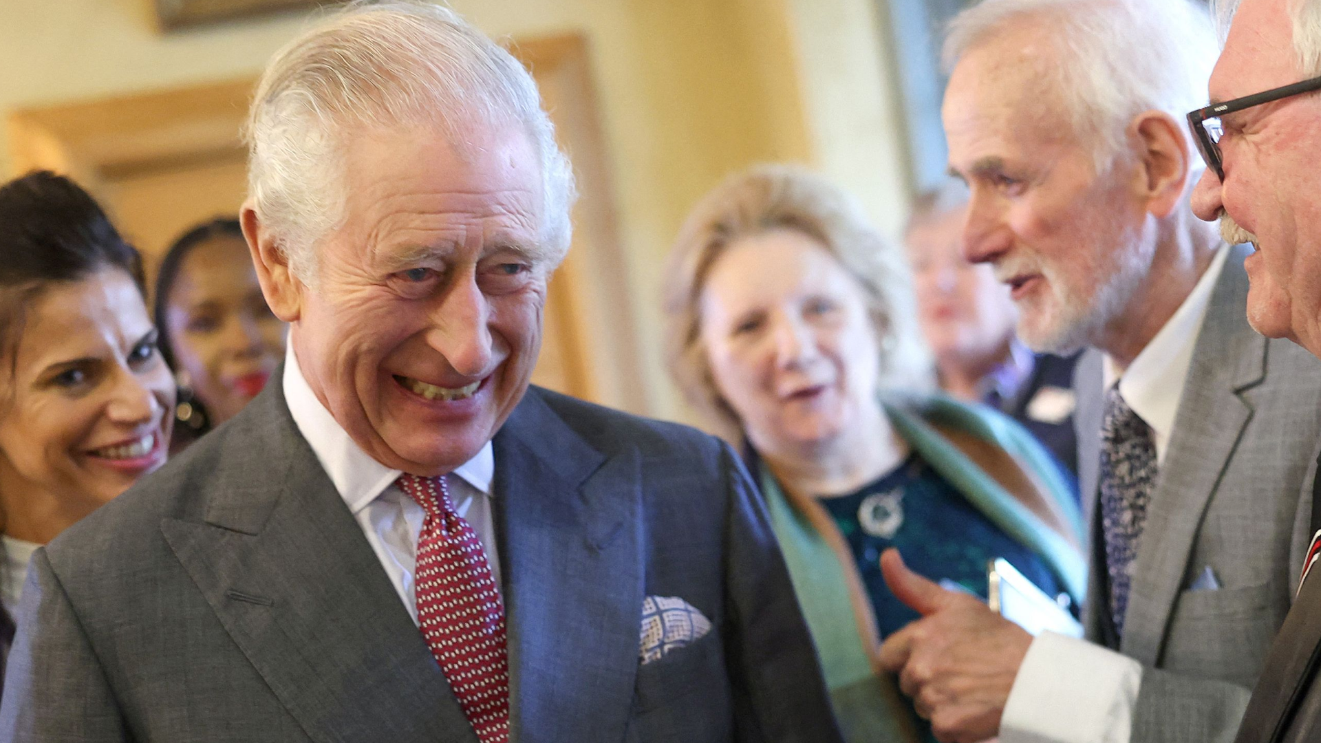 King Charles laughing in a gray suit and red tie