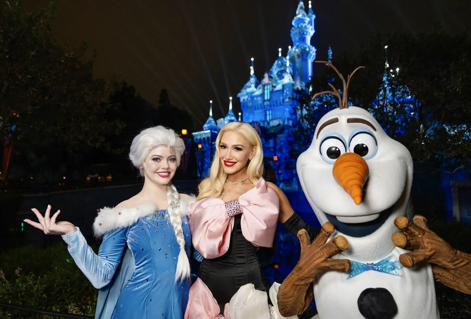 gwen stefani wears a dress with big pink bows and poses with elsa and the snowman from frozen in front of the disney castle in a promo shot for the wonderful world of disney holiday spectacular