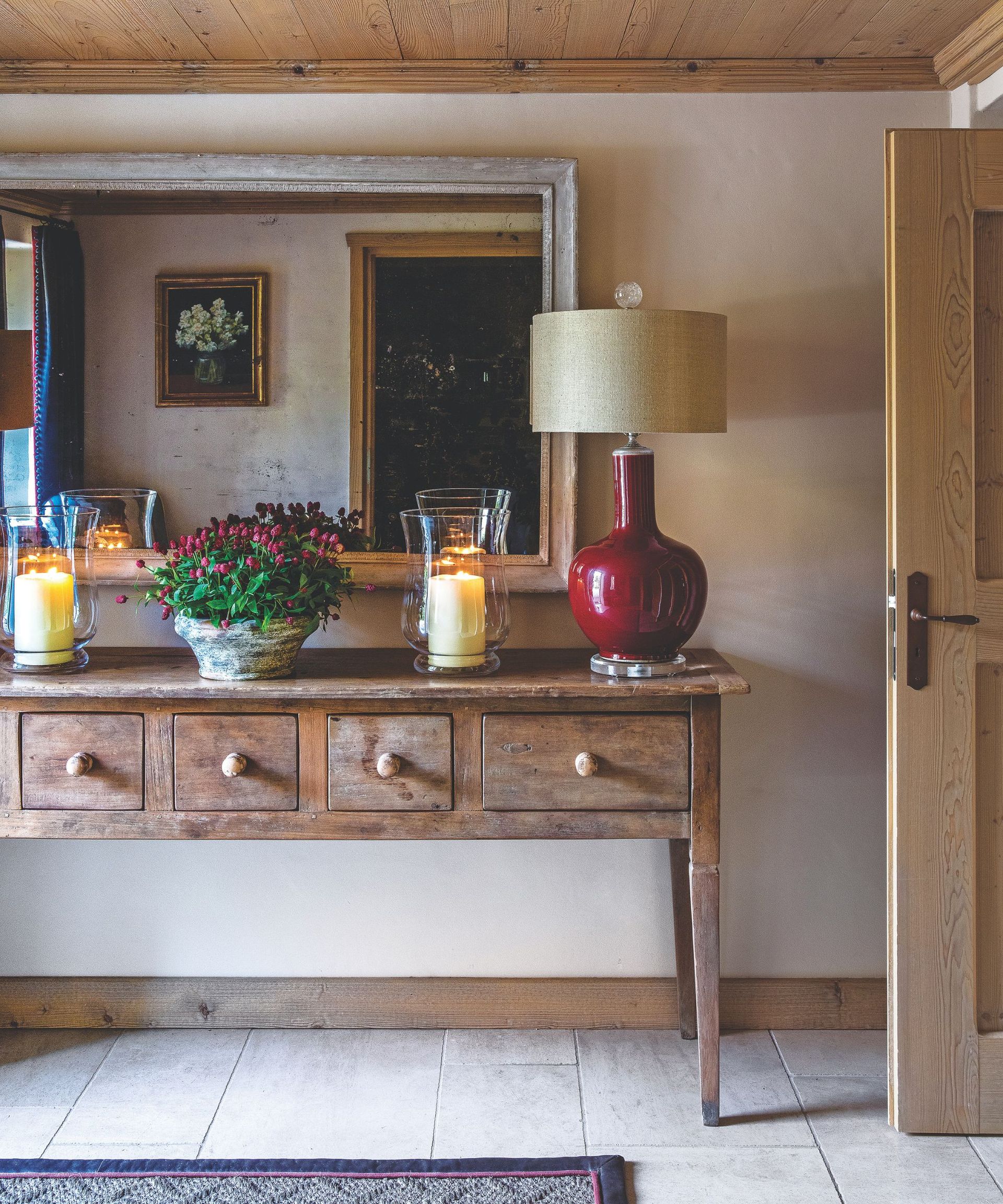 Wooden console table in hallway with large red lamp on and two glass storm lanterns with lit candles and a bowl of flowers in beween; country rustic hallway