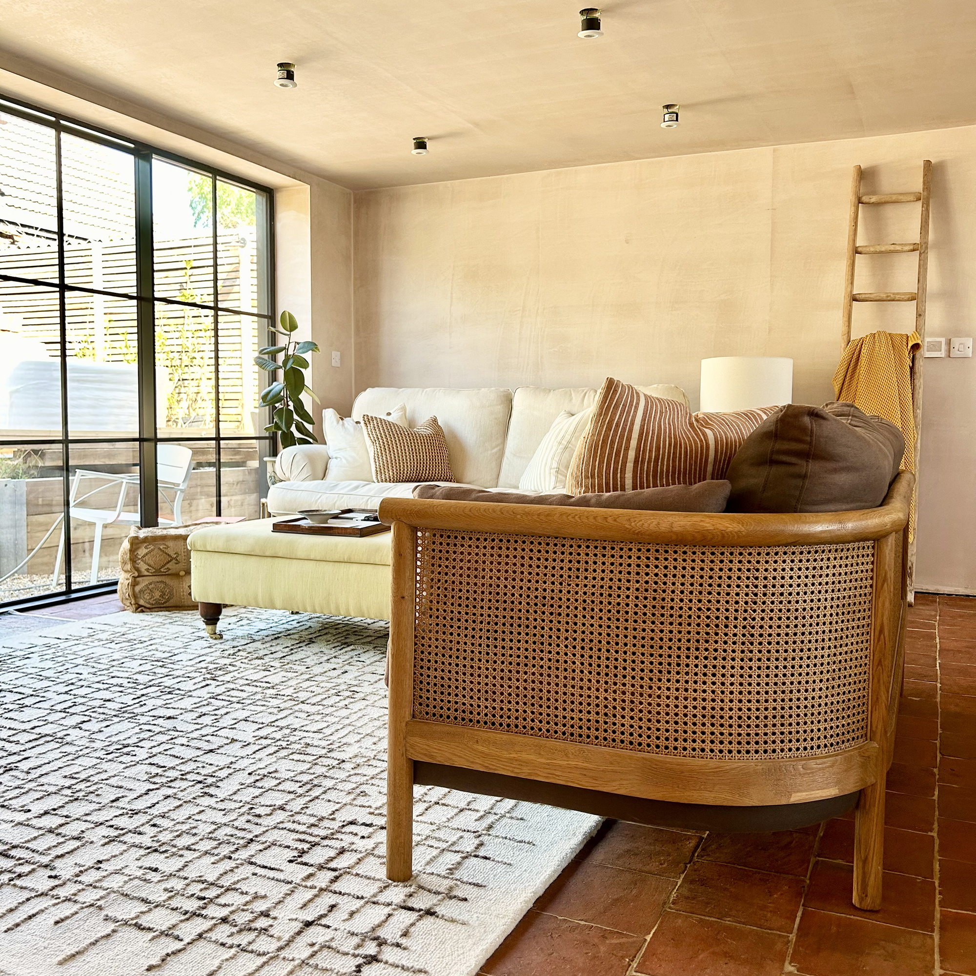 neutral living room with rattan sofa, terracotta floor tiles and a large neutral rug. Black framed sliding doors open up to the garden