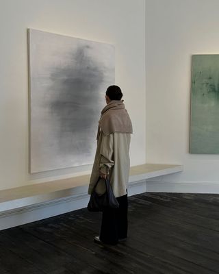 Jordan Risa wearing a trench coat, black bag, and a camel sweater draped over her shoulders in a museum.