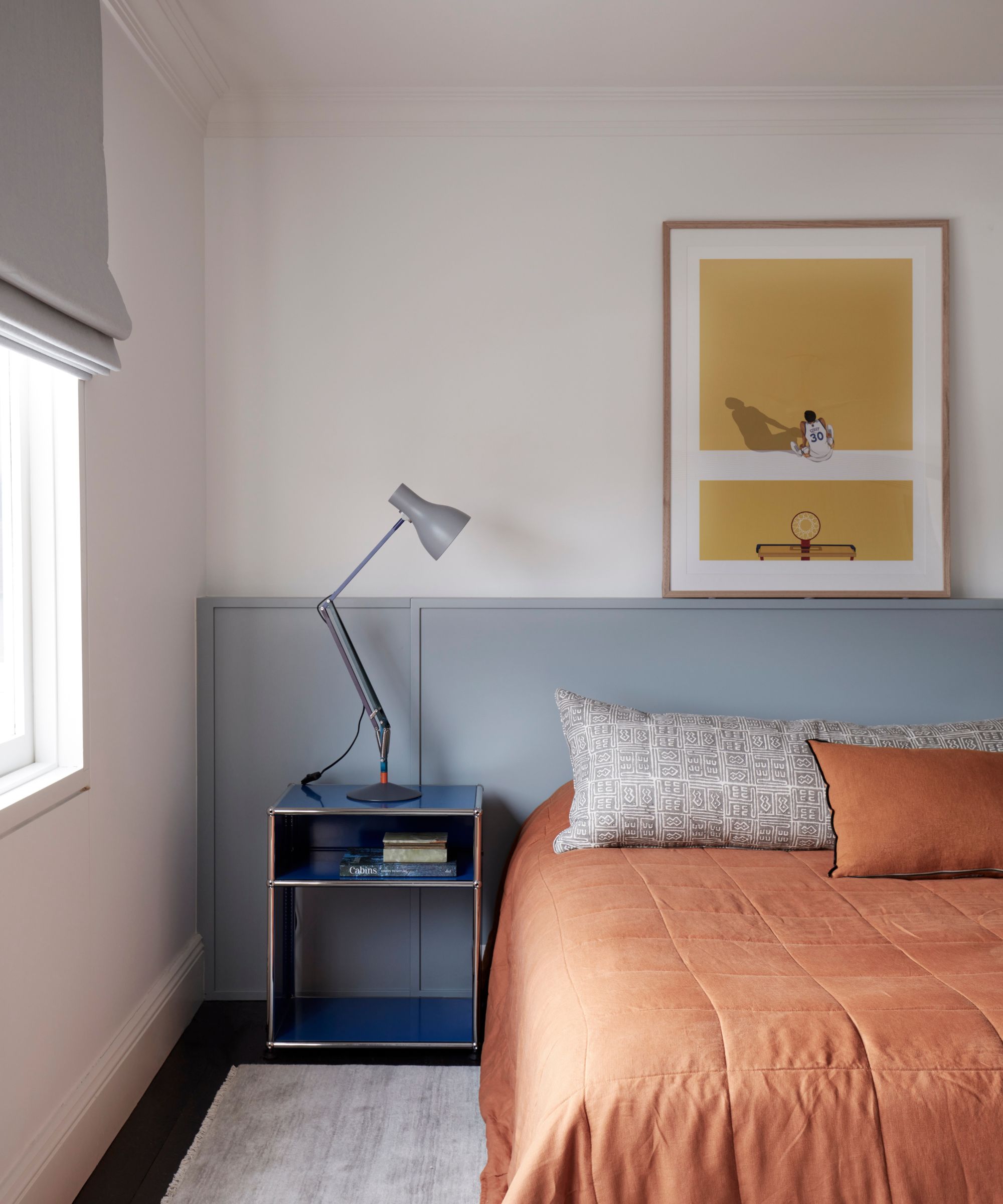 Bedroom with pale blue painted ledge holding yellow framed print, low bed with orange quilt, blue nightstand and angle poise lamp