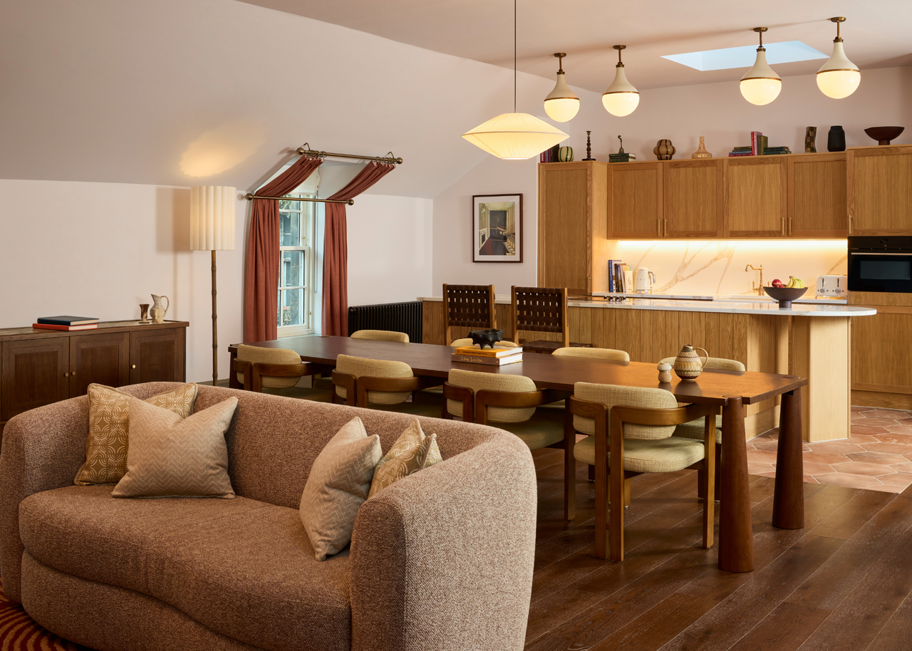 A bright living room with overhead lighting, a brown couch, a long wood dining table with three-legged dining chairs, and a kitchen island with woven leather stools, and a backlit stone backsplash by wood kitchen cabinets
