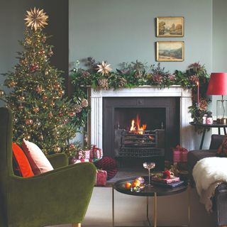 A sage green-painted living room with a lit Christmas-decorated fireplace and Christmas tree covered in lights