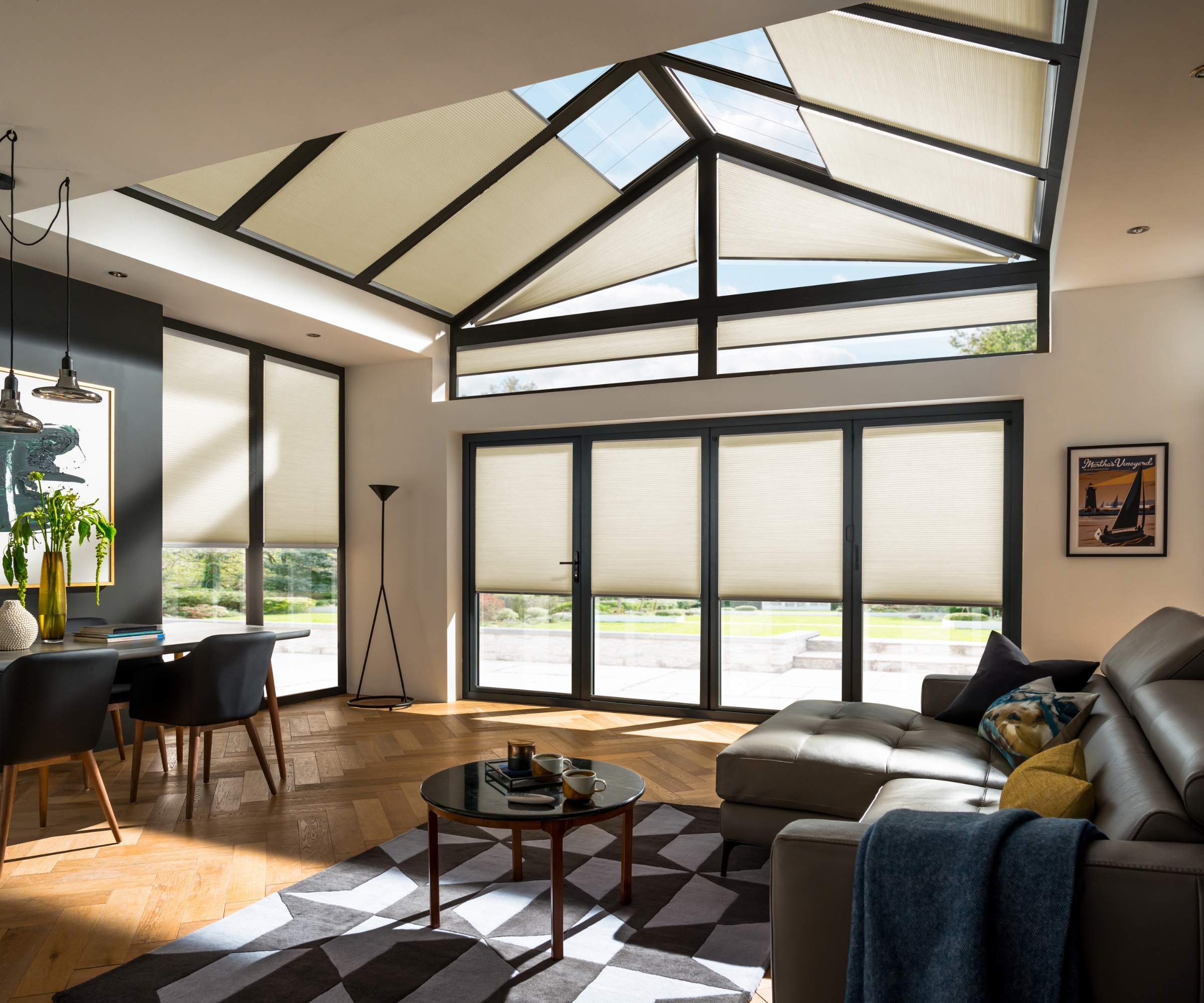 Vaulted glass roof with cream cellular blinds over dining table and dark leather sofa, patterned rug.