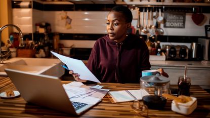 A woman looks at paperwork while sitting with her laptop at the kitchen counter.