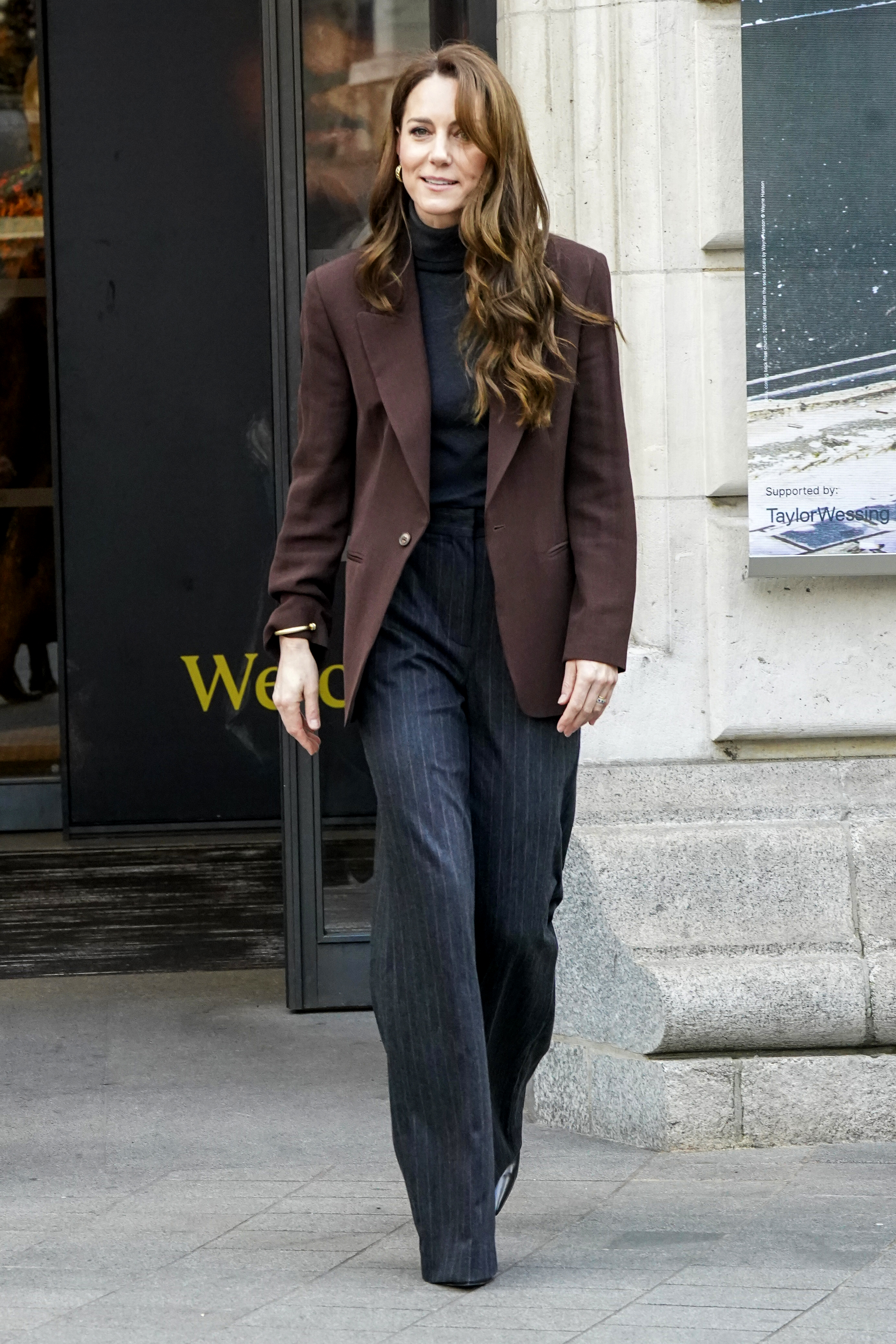 Kate Middleton has long brown wavy hair and wears a brown blazer with jeans, while wearing a tartan overcoat and heels