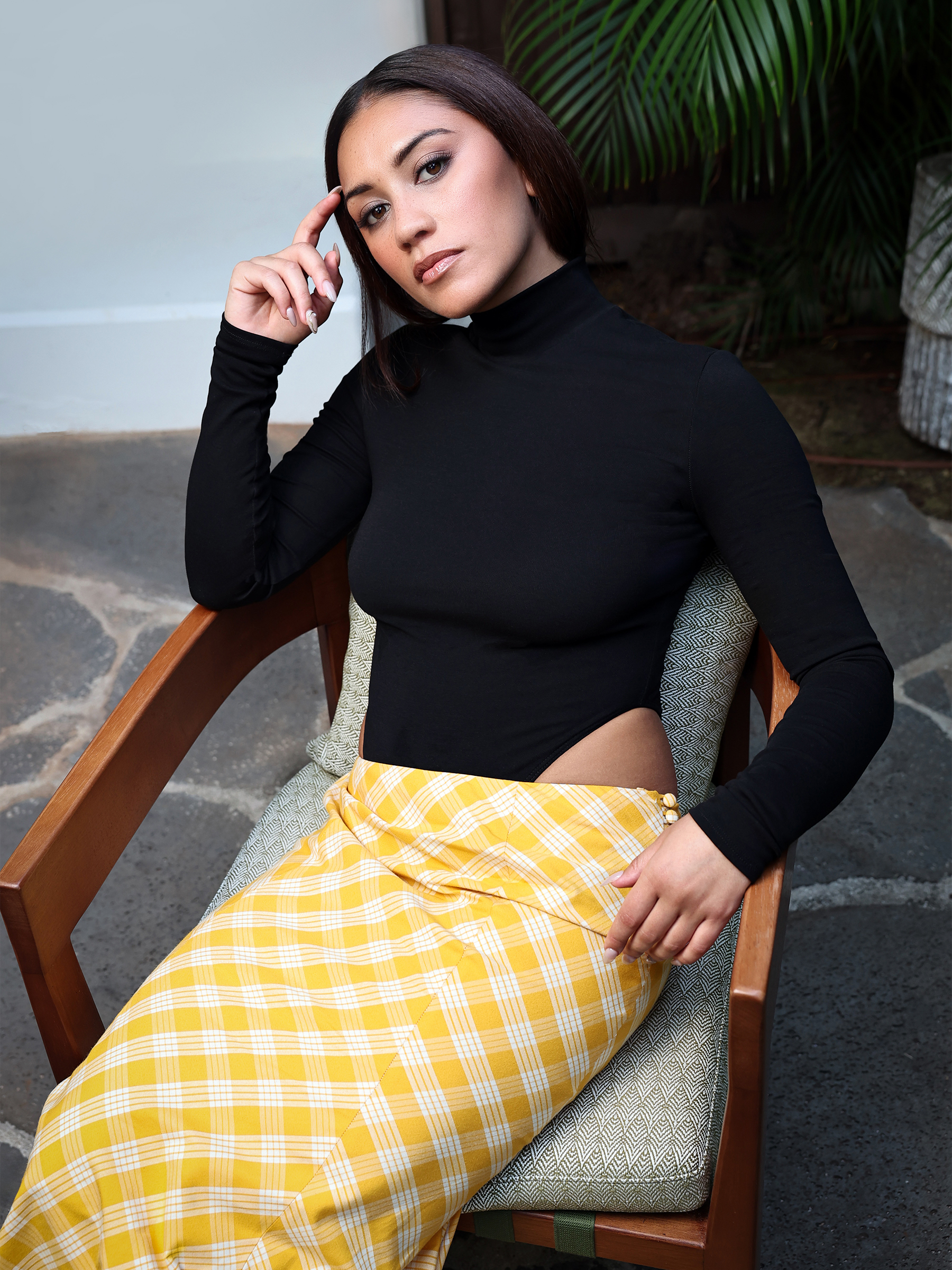Luciane Buchanan photo taken during a press day for her new show Chief of War. She is wearing a custom look by Hawaiian designer Rocket Ahuna consisting of a black high neck, long-sleeve top that cuts up on the side and a yellow and white gingham maxi skirt.