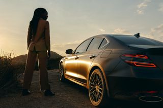 Genesis G70 car exterior image at sunset with woman standing to left of vehicle