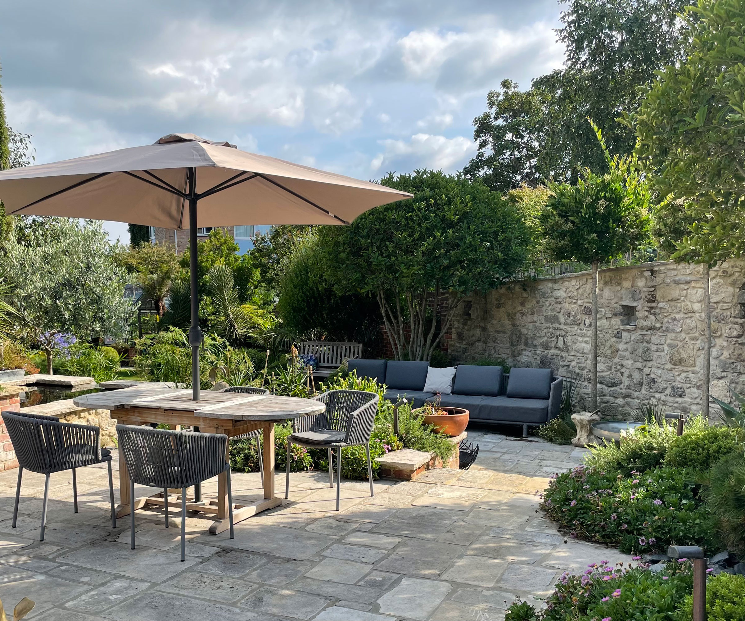 enclosed courtyard garden with brick wall, paving and garden furniture including dining set, parasol and sofas, with planting beds and multi-stemmed evergreen tree
