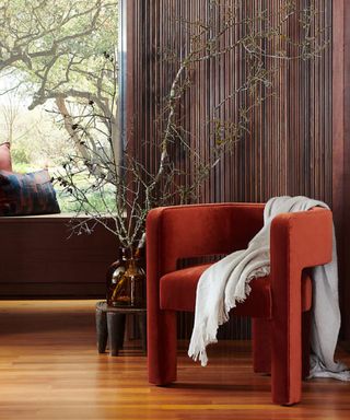 Dark orange velvet sculpted chair with rounded back and arms, with white throw against a dark wood paneled wall and floor to ceiling window