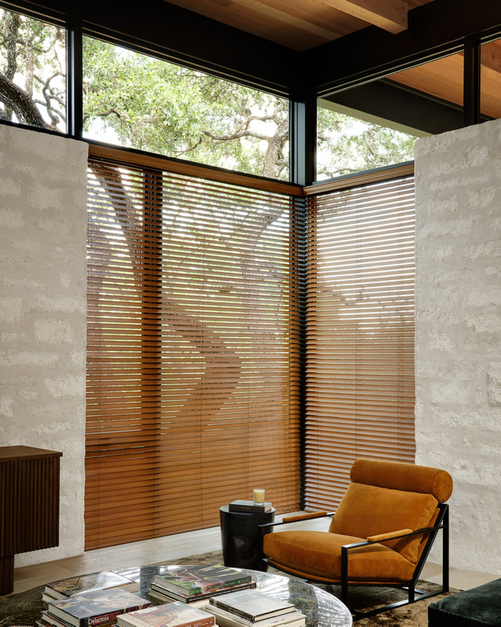 modern living room with stonewashed walls, brown timber venetian blinds, a timber ceiling, high windows, a round coffee table covered in books, a statement rust-colored velvet armchair and a side table