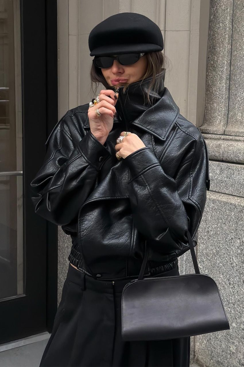 A woman standing outside wearing a black leather funnel-neck jacket, black wide-leg trousers, a black bowler bag, black baker cap, and stacked gold and silver rings.
