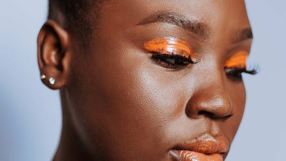 A woman with glowing skin and bright orange eyeshadow is pictured against a blue background to really make the contrasting eyeshadow pop