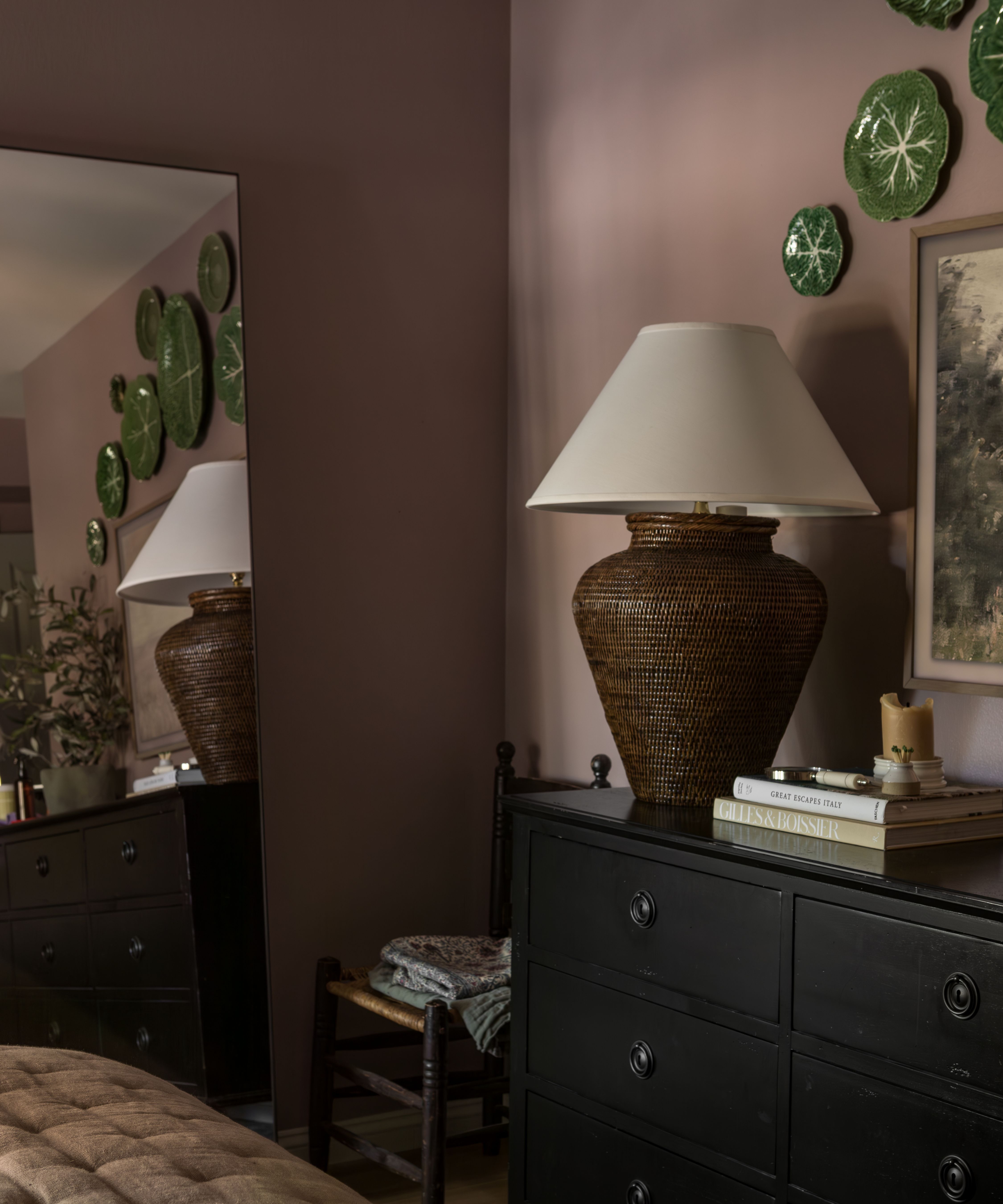 a main bedroom painted in sulking room pink with a black dresser, floor length mirror, rattan table lamp and cabbage plates on the wall