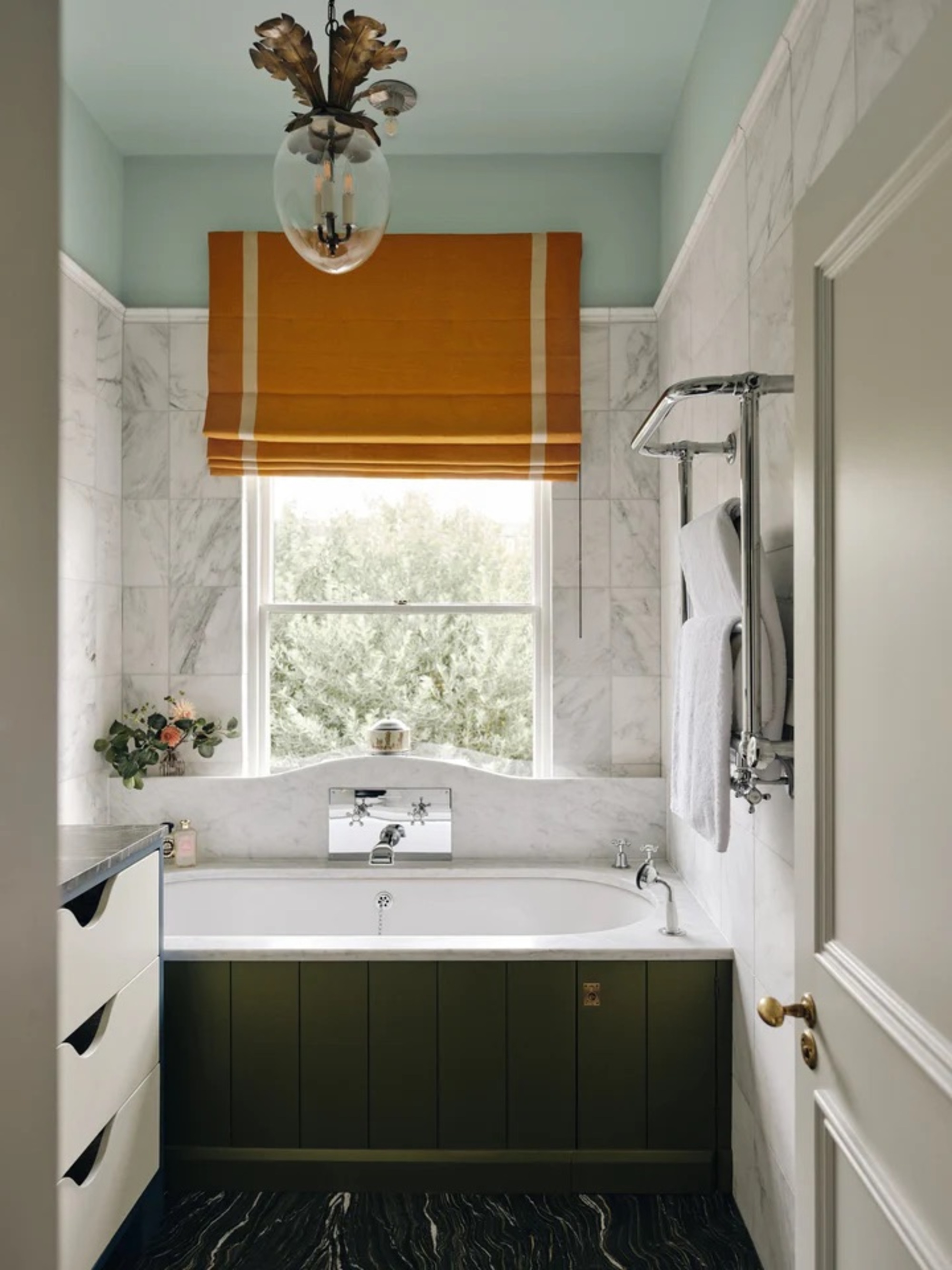 small colorful bathroom with light blue ceiling, mustard yellow roman blind over window above built-in bathtub painted green with black floors and marble wall tiles