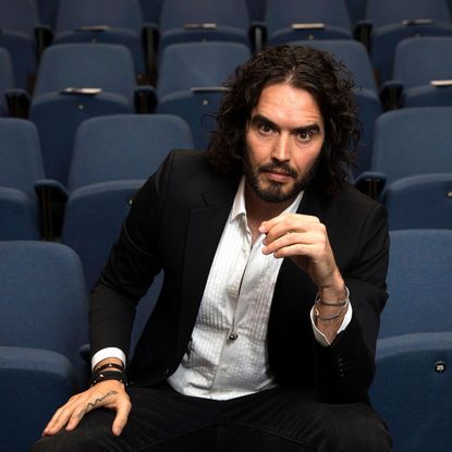 Comedian Russell Brand poses in a suit seated in a theatre seating area
