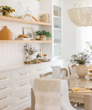 white dining room with shiplap above cabinets, open shelving, chandelier, table to right, chairs, light and airy