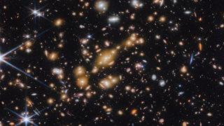  A field of galaxies on the black background of space. In the middle is a collection of dozens of yellowish galaxies that form a foreground galaxy cluster. Among them are distorted linear features, which mostly appear to follow invisible concentric circles curving around the centre of the image. The linear features are created when the light of a background galaxy is bent and magnified through gravitational lensing. A variety of brightly coloured, red and blue galaxies of various shapes are scattered across the image, making it feel densely populated