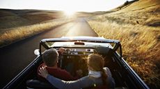 An older couple drive toward the sunset in a convertible out in the country.