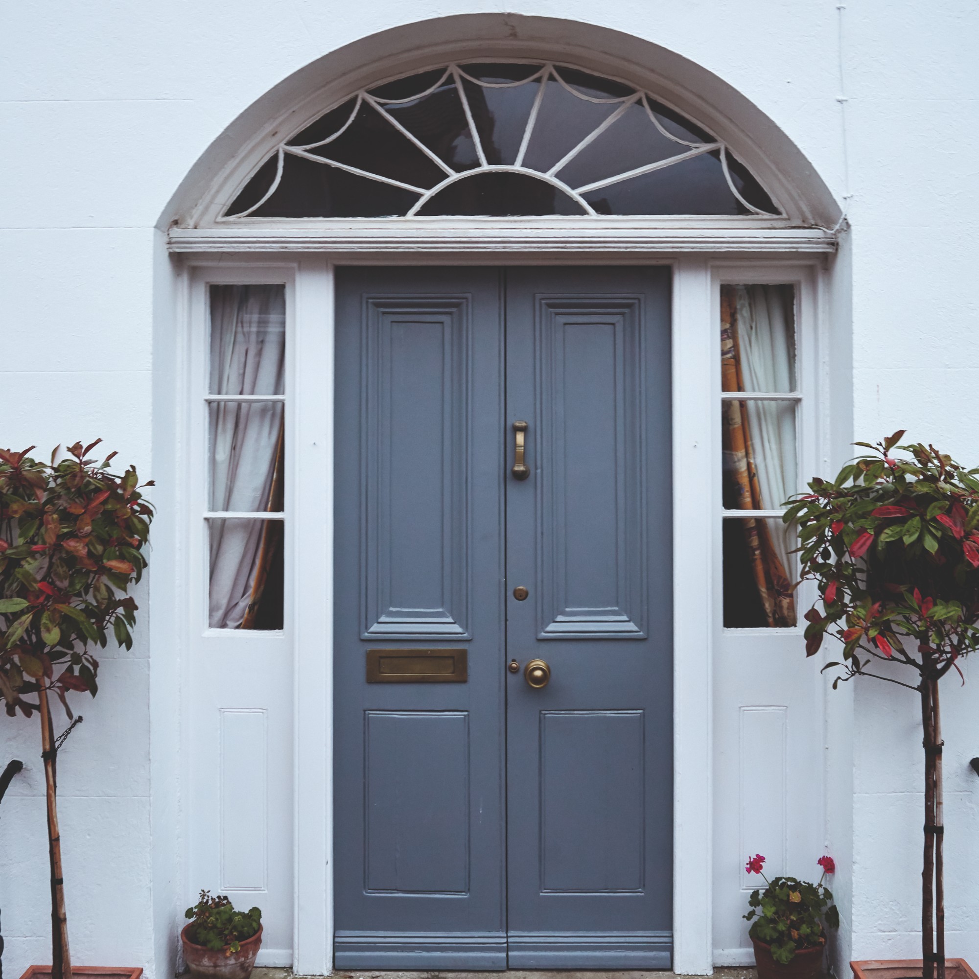 A white house exterior with a grey-painted double door with decorative potted trees on either side