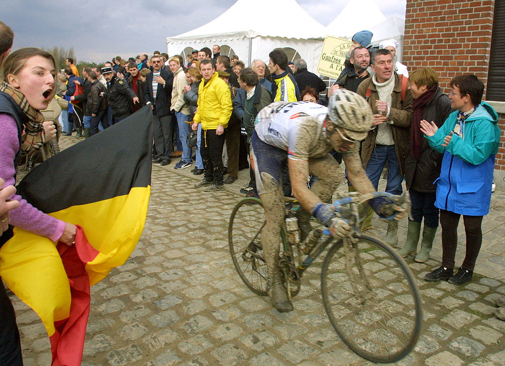 Foto lateral de Servais Knaven, cubierto de barro, en un tramo adoquinado de la París-Roubaix 2001, lleno de seguidores