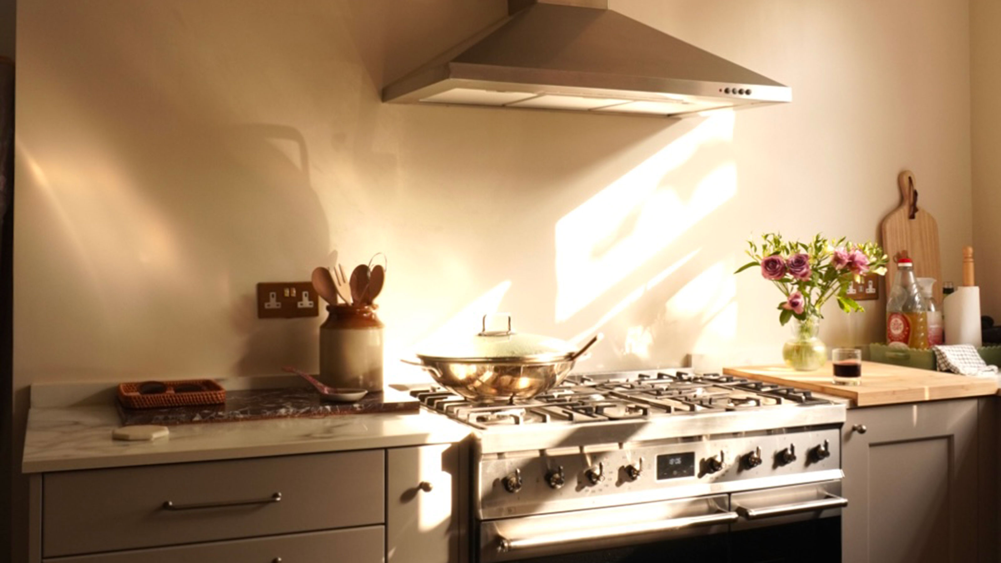 Cream kitchen with stainless steel stove and extractor fan, framed by ceramic vase with wooden spoons, vase of flowers and wooden serving boards