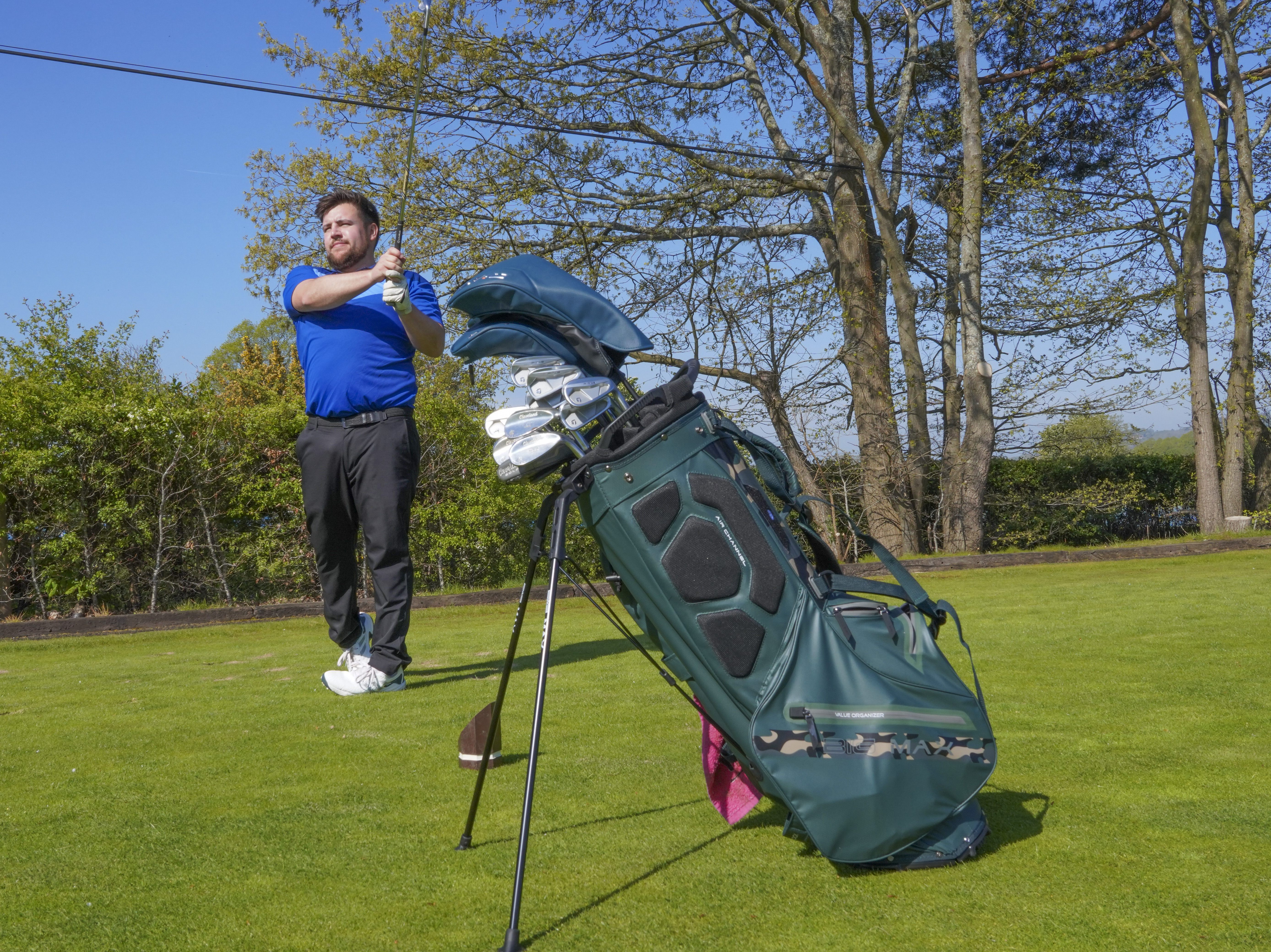 A golfer hits a golf ball, with the Big Max Aqua Eight H Stand Bag in front