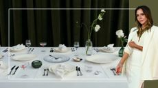The Victoria Beckham x Westwing laid on a white table with the hammered cutlery, tealight holder, butter dishes, and bud vases, next to a cutout of Victoria Beckham