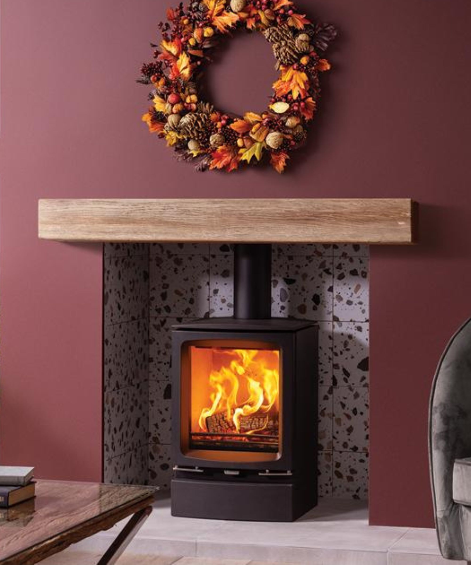 A modern black log burner with vivid flames is set against a terrazzo-tiled fireplace surround. A light wood mantel rests above, with an autumnal wreath of leaves, berries, and pinecones hanging on the wall above it.