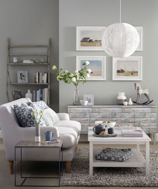 grey living room with a wooden book shelf and a chest drawer topped with flowers and accessories, and a white sofa with blue patterned cushions and a white wooden coffee table on top of a furry grey carpet