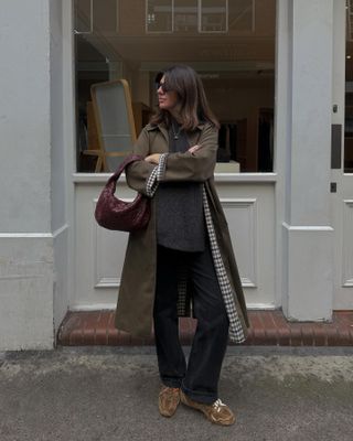 style influencer Francesca Saffari wearing a gingham print lined trench coat, gray sweater, burgundy Bottege Veneta bag, dark cuffed jeans, and New Balance x Miu Miu brown suede sneakers