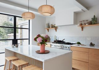 A marble topped island and timber cabinets opposite it. On the island is a vase with flowers inside of it. Above the island are two bamboo pendant lights hanging. There is also open shelving above the countertops with daily essentials.