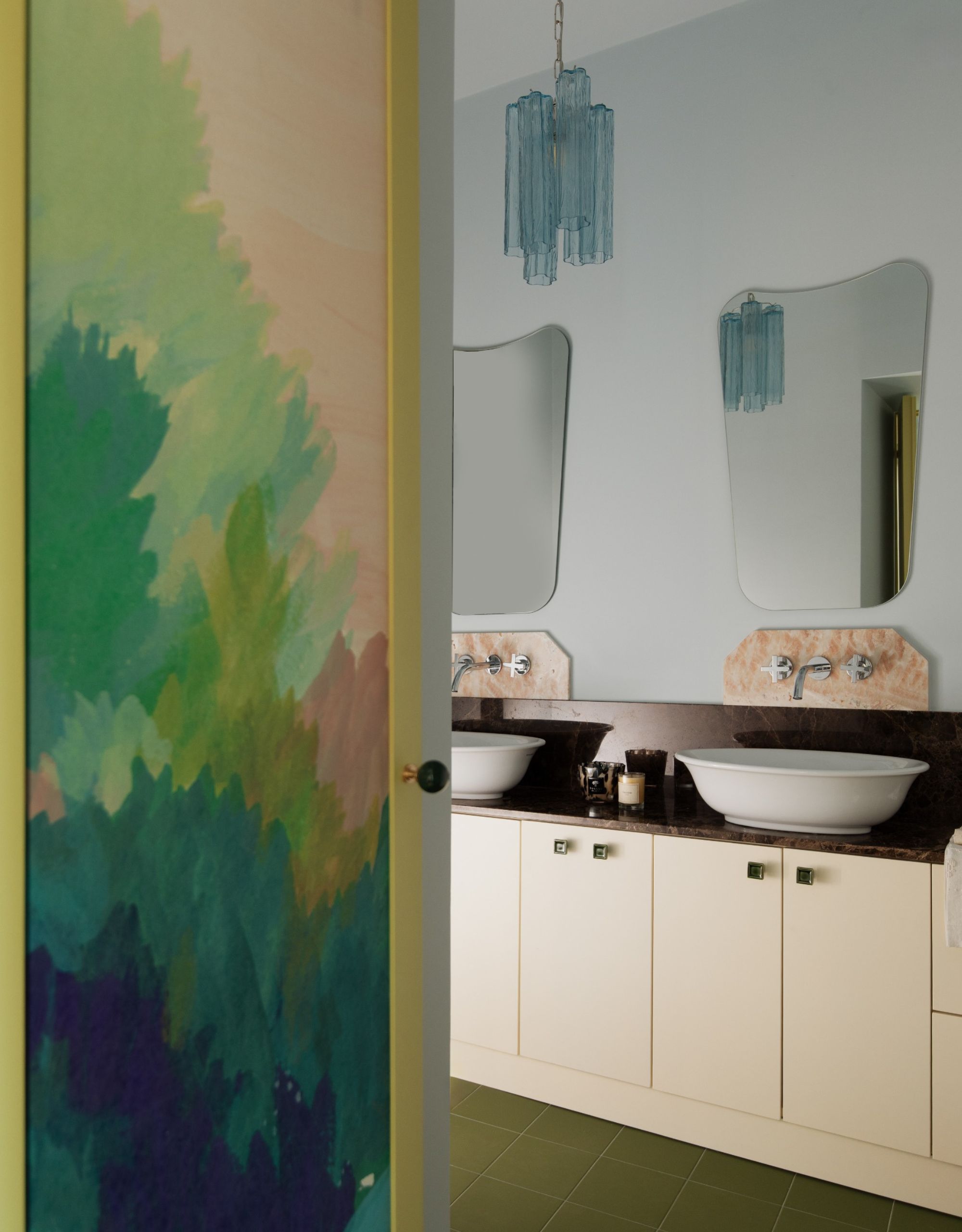 A double basin vanity in a bathroom with a wall mirror above each sink and a blue murano-glass style chandelier hangs in between the mirrors with a mural-painted doorway leading to the space