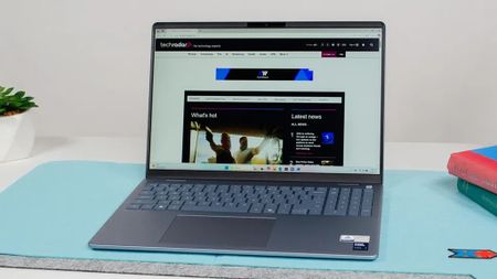 A sleek, open laptop on a light blue desk mat displays a tech website. A small plant and books are in the background, conveying a clean, modern workspace.