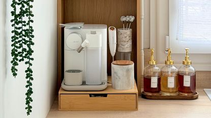 Close-up image of a white coffee machine on a wooden stand in a white kitchen. There is a tray of three syrups beside it and other coffee accessories.