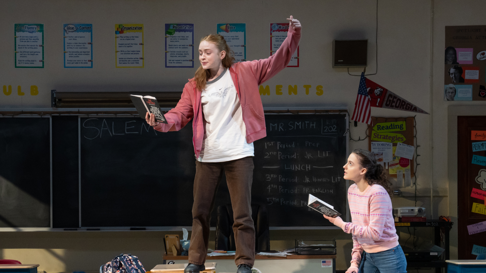 sadie sink stands on a table holding a book while another girl looks on in a shot of the play john proctor is the villain