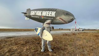 Man in shark costume larks about in front of the Shark Week 2022 blimp