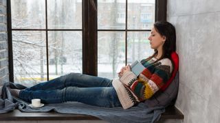 A woman sleeping in a windowsill wearing a knitted jumper with snow falling outside