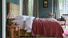 deep earthy blue bedroom styled with a yellow tartan upholstered bed, white linens, a rust red throw and a rattan side table with pleated table lamp