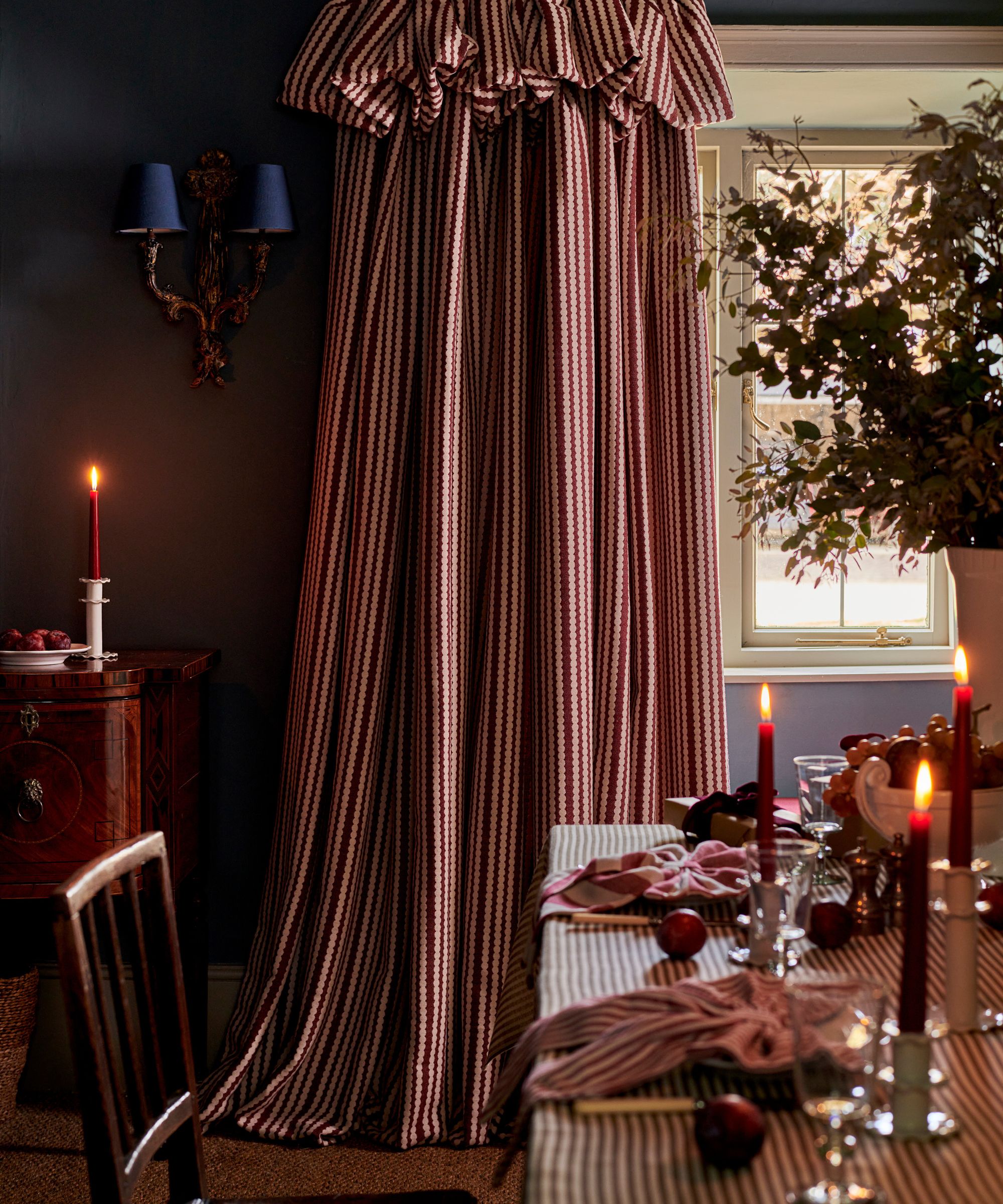 A cozy dining room with a festive Christmas tablescape including a red striped tablecloth, napkins, and red taper candles. Long red striped curtains cover the window in the corner of the room.