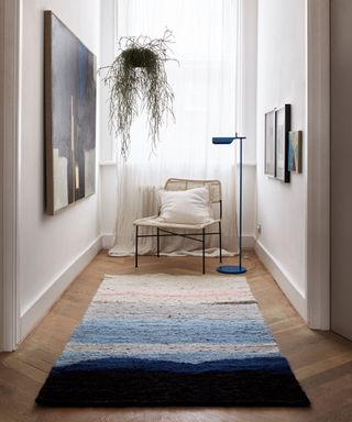 Wooden floored hallway with a white curtained window, chair and cushion with a reading lamp, wall hung artwork and hanging plant basket.