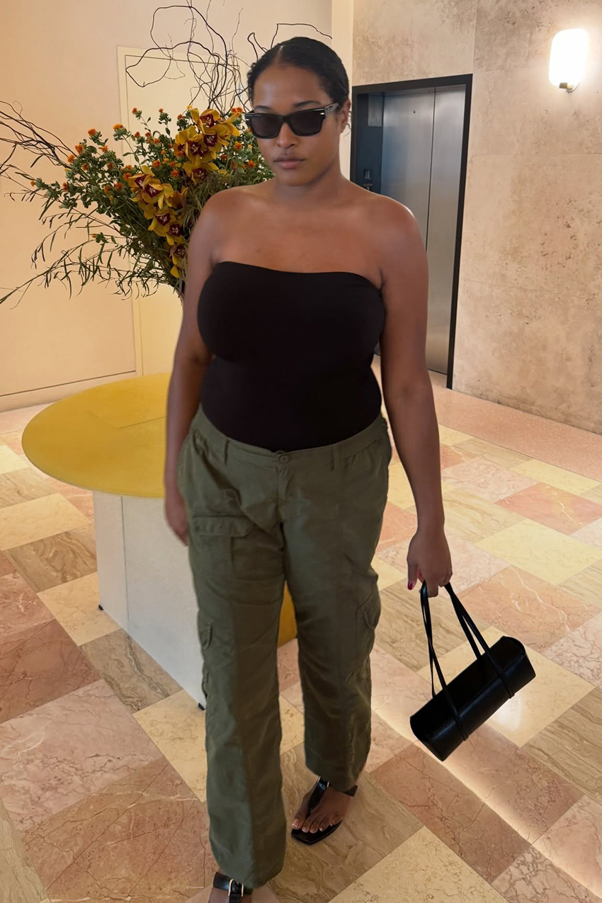 A woman is wearing a black tube top, green cargo pants, black sandals, and a black leather East West bag