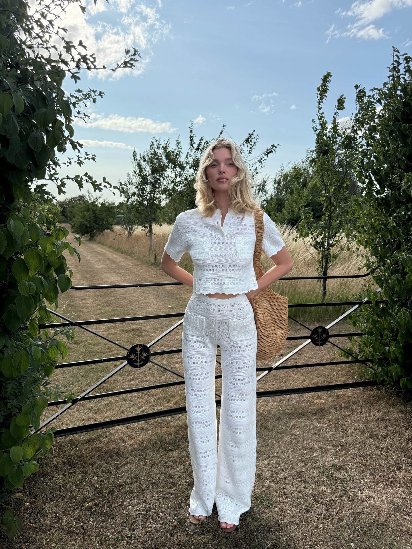 Elsa Hosk stands outside wearing a white crochet top and trouser co-ord with a brown woven bag and brown sandals.