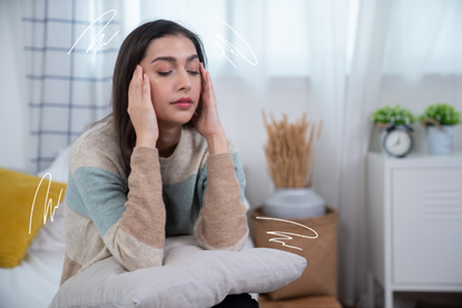 Woman experiencing a migraine headache in her bedroom with pillow on her lap and curtains closed behind her