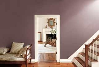 Purple-painted hallway with the start of a staircase in view and a stripped wooden bench. There is an open door frame in the middle leading to a traditional white living room